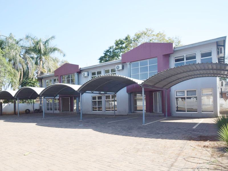 Front view of a commercial office building featuring a paved parking area with covered carports and multiple aluminium-framed windows.