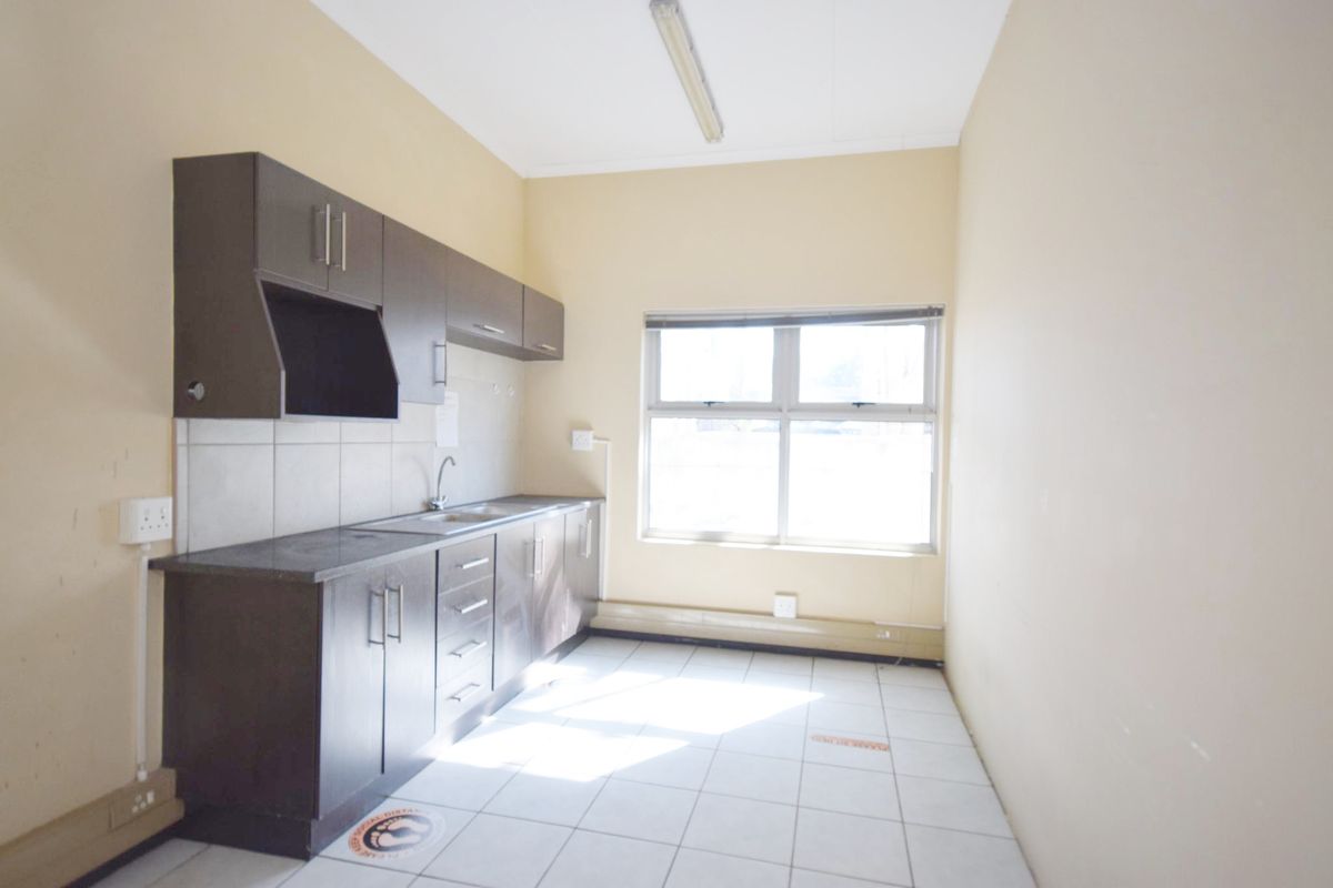 A functional office kitchenette featuring dark built-in cabinetry, laminate countertops, a stainless steel sink, and tiled flooring in a neutral workspace.