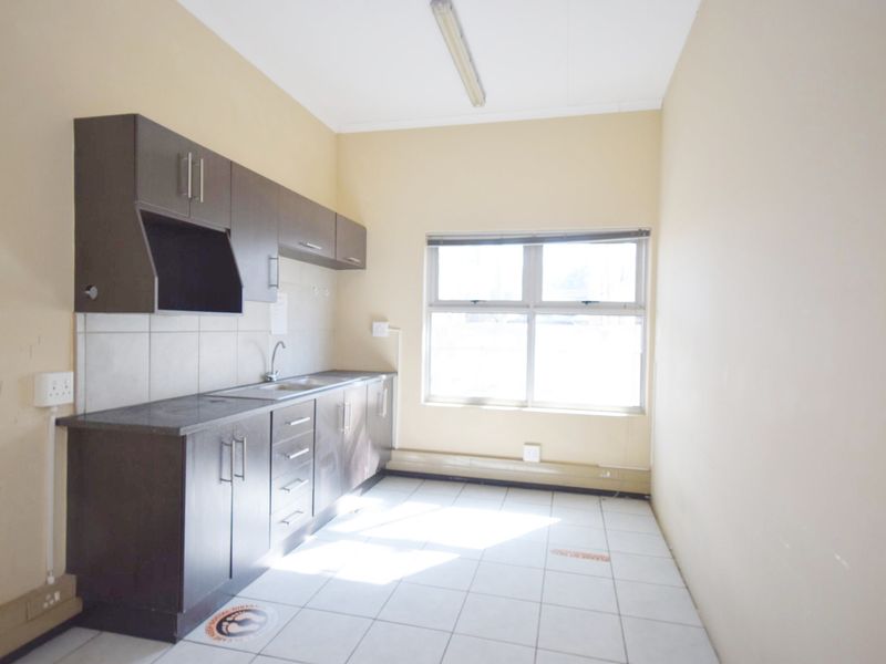 A functional office kitchenette featuring dark built-in cabinetry, laminate countertops, a stainless steel sink, and tiled flooring in a neutral workspace.