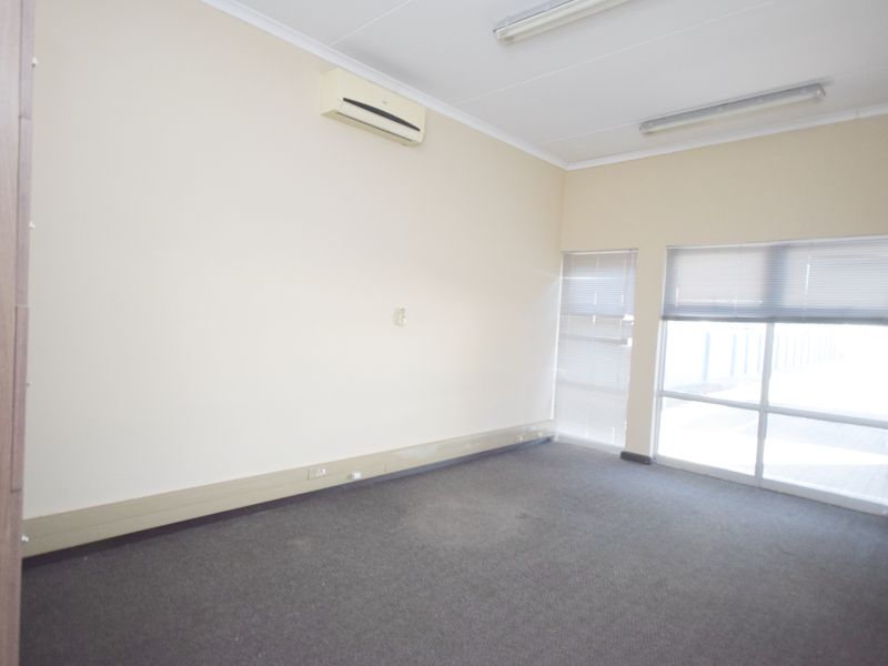 A vacant office space featuring neutral walls, carpet flooring, large windows with venetian blinds, and a wall-mounted air conditioning unit.