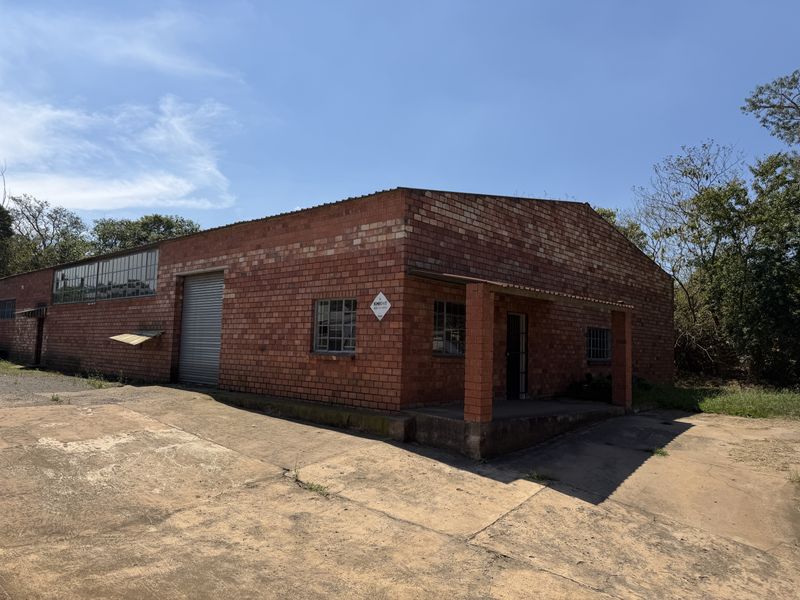 Industrial warehouse building featuring a red brick facade, roller shutter door, steel-framed windows with security bars, and a concrete apron entrance area.