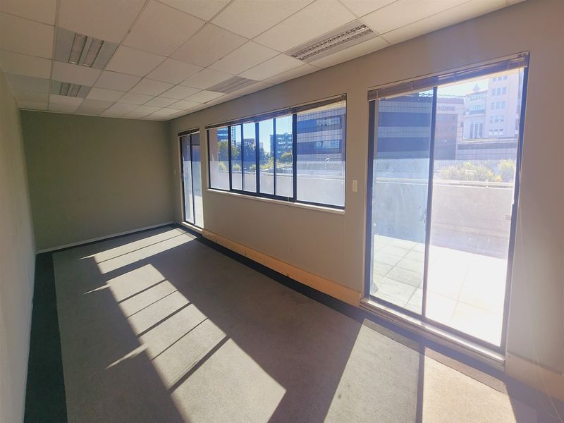 A bright, empty office space featuring a suspended ceiling, carpeted flooring, and large sliding glass doors providing access to an outdoor balcony area.