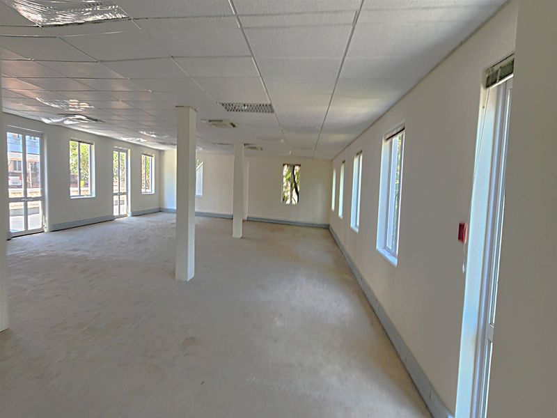 A spacious, open-plan office area featuring a suspended ceiling, structural columns, and multiple windows providing ample natural light.