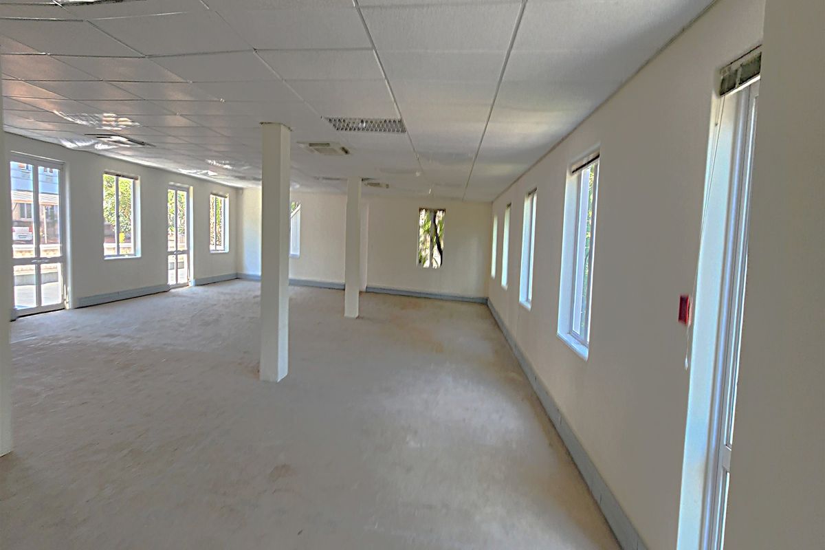 A spacious, open-plan office area featuring a suspended ceiling, structural columns, and multiple windows providing ample natural light.