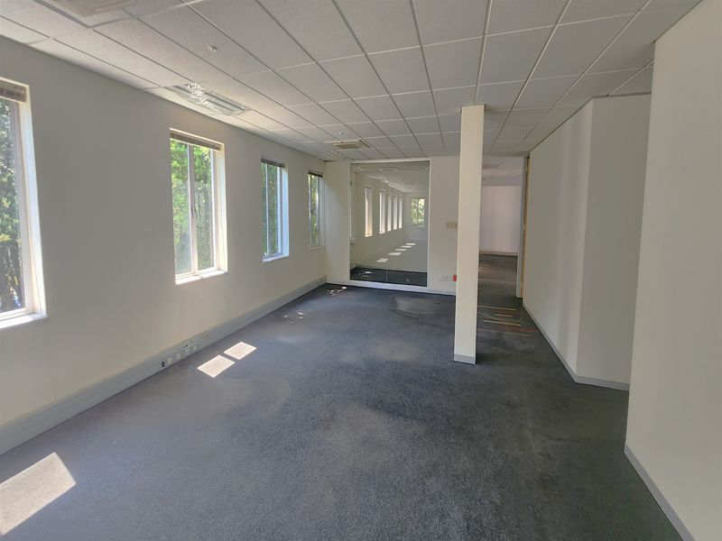 An empty, open-plan office space featuring a suspended ceiling, carpeted flooring, large windows for natural light, and a central structural column.