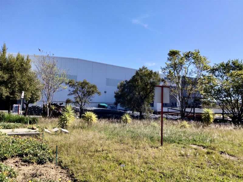 Exterior view of an industrial warehouse facility featuring metal cladding, set behind an overgrown landscaped area.