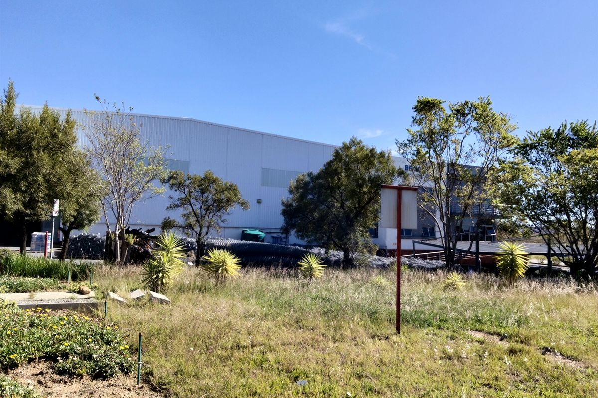 Exterior view of an industrial warehouse facility featuring metal cladding, set behind an overgrown landscaped area.