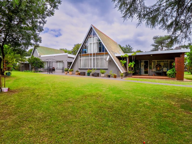 Rear view of a unique A-frame style home featuring a spacious lawn, mature trees, and a covered patio area.