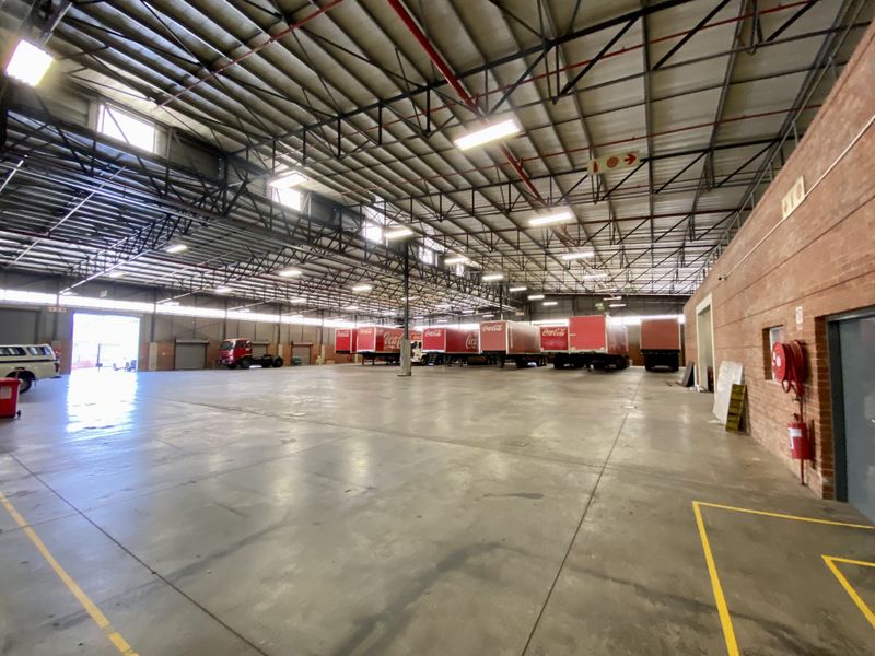 Spacious industrial warehouse featuring high ceilings with exposed steel trusses, concrete flooring, integrated sprinkler system, and multiple roller shutter access points.