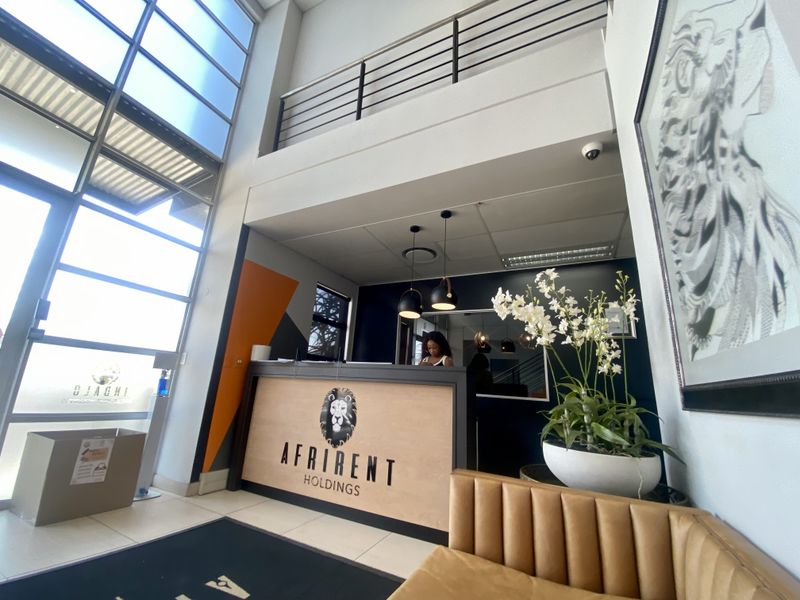 Modern industrial reception area featuring double volume ceilings, a mezzanine level with metal balustrades, and a large glass facade providing ample natural light.