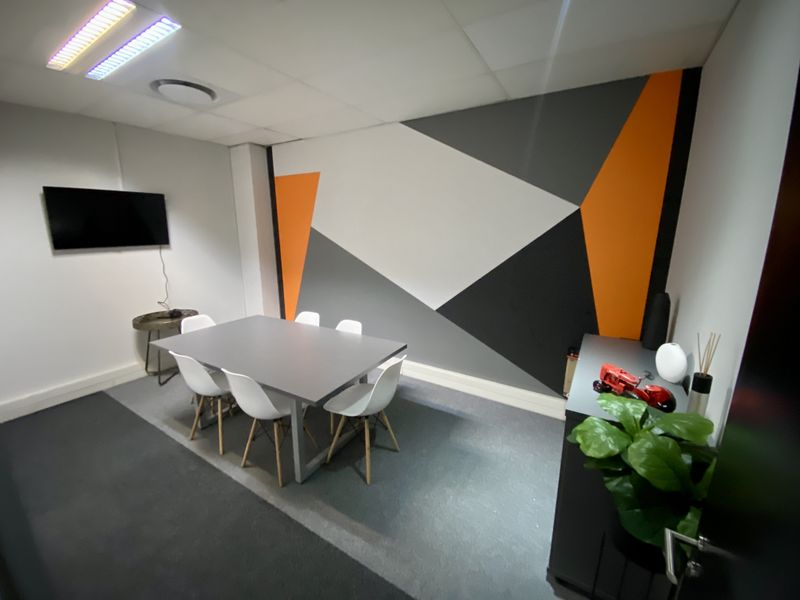 A modern office meeting room featuring a geometric accent wall, suspended ceiling with recessed lighting, and grey carpeted flooring.