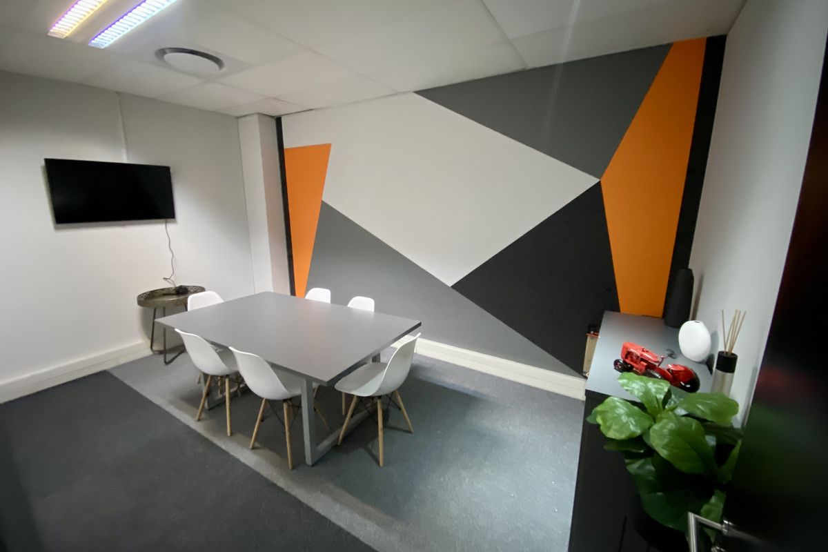 A modern office meeting room featuring a geometric accent wall, suspended ceiling with recessed lighting, and grey carpeted flooring.