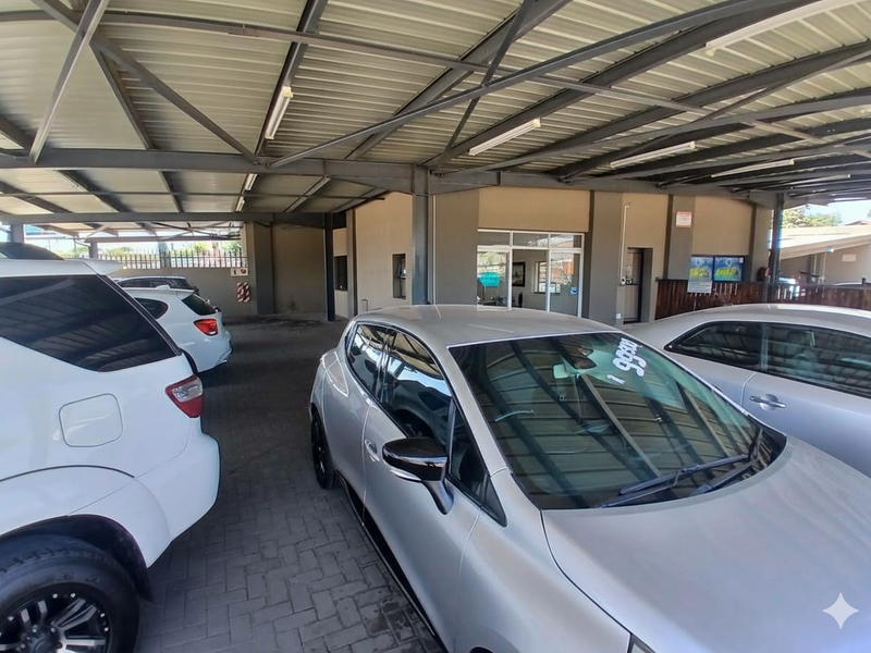 A covered commercial parking area featuring a steel roof structure, paved flooring, and fluorescent lighting, providing sheltered access to the building entrance.