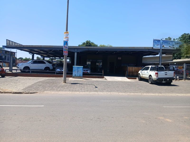 Front exterior view of a commercial property featuring a large steel canopy structure, paved driveway, and perimeter metal fencing.