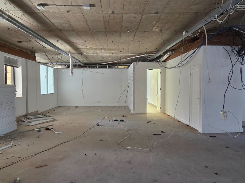An open-plan office space currently under renovation, featuring an exposed concrete ceiling, unfinished flooring, and exposed electrical infrastructure.