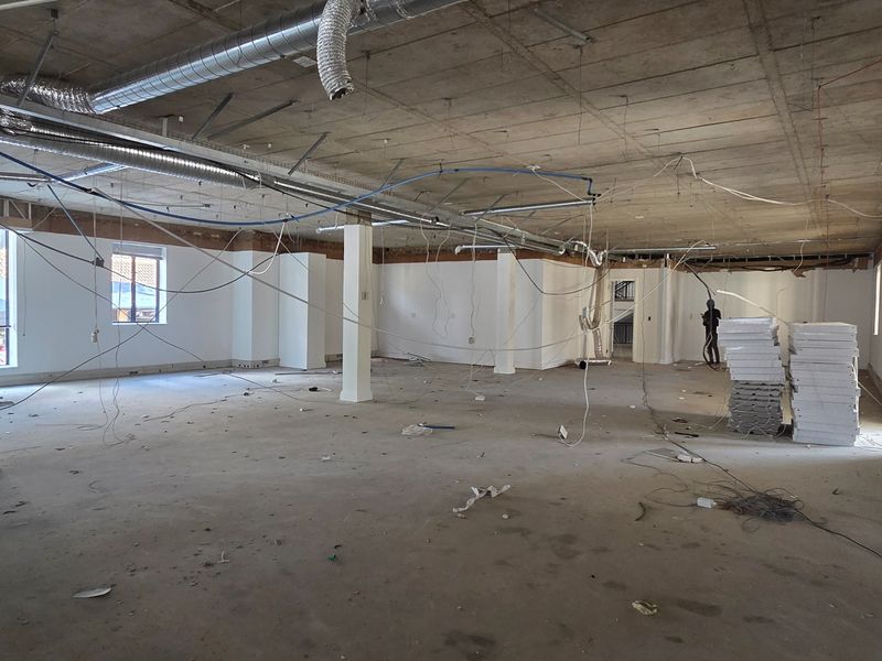 A large, open-plan office space currently under renovation, featuring exposed concrete ceilings, structural columns, and unfinished electrical and HVAC systems.