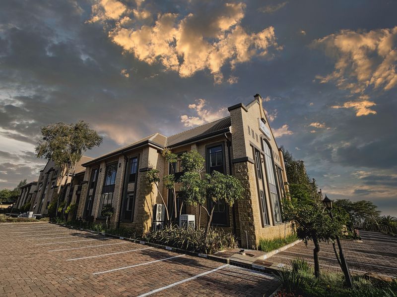 A professional exterior view of a multi-story office building featuring a brick facade, tiled roof, and a paved parking area with landscaped surroundings.