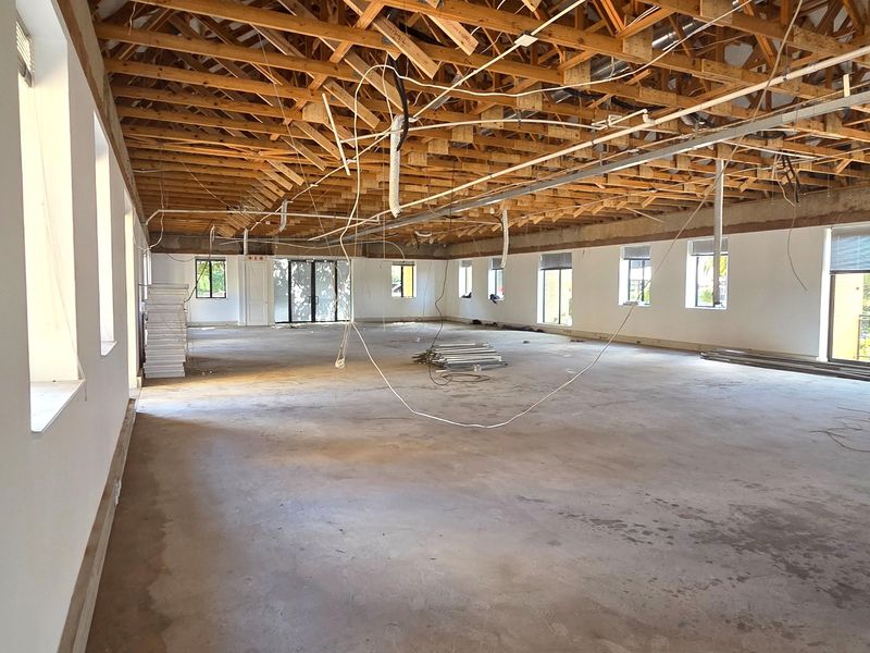 A large, open-plan office space currently under renovation, featuring exposed timber roof trusses, concrete flooring, and large windows providing ample natural light.