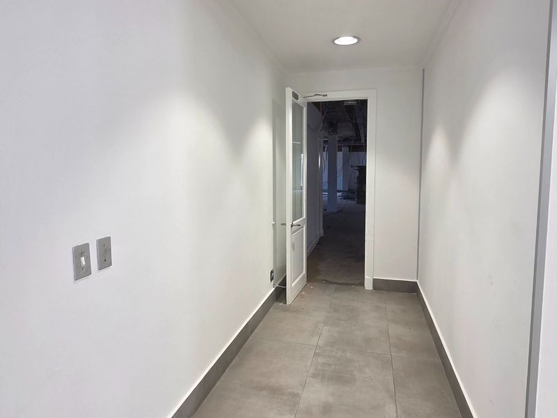 A clean, minimalist hallway featuring grey tiled flooring, recessed lighting, and a glass-panelled door leading into an unfinished office space.