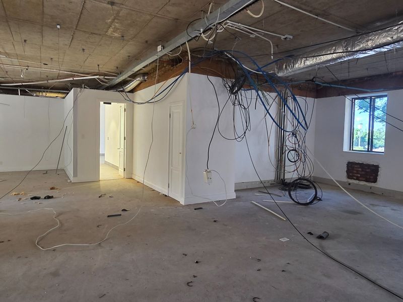 An unfinished office space featuring exposed concrete ceilings, exposed electrical and HVAC infrastructure, and a large window, requiring significant renovation to complete.