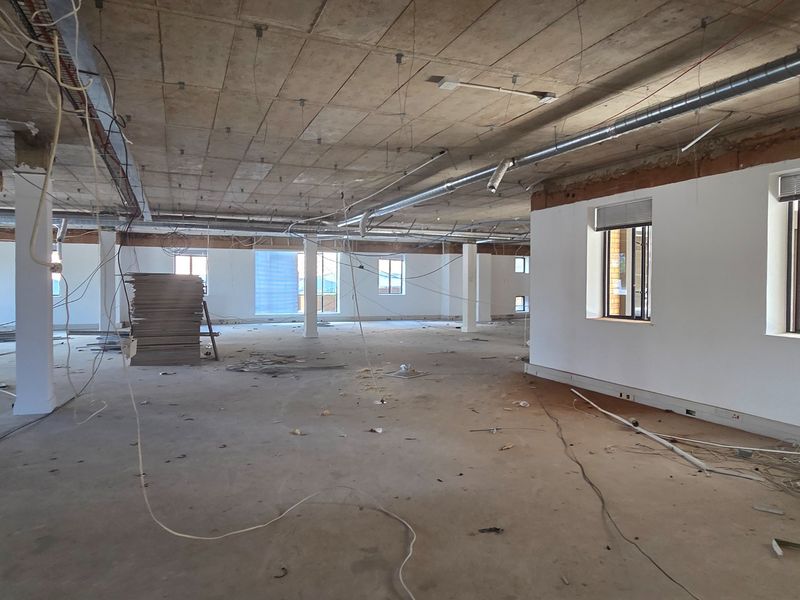 A large, open-plan office space currently under renovation, featuring exposed concrete ceilings, industrial ducting, and large windows providing ample natural light.