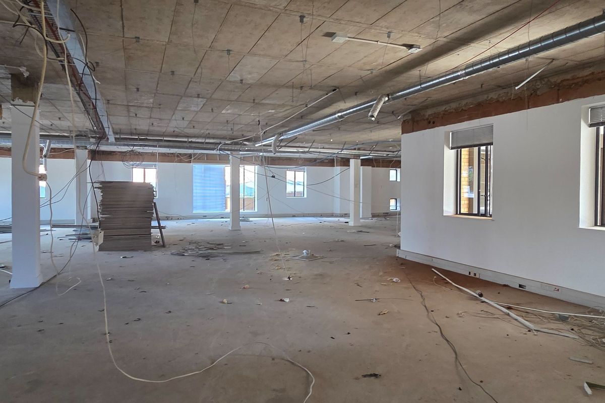 A large, open-plan office space currently under renovation, featuring exposed concrete ceilings, industrial ducting, and large windows providing ample natural light.