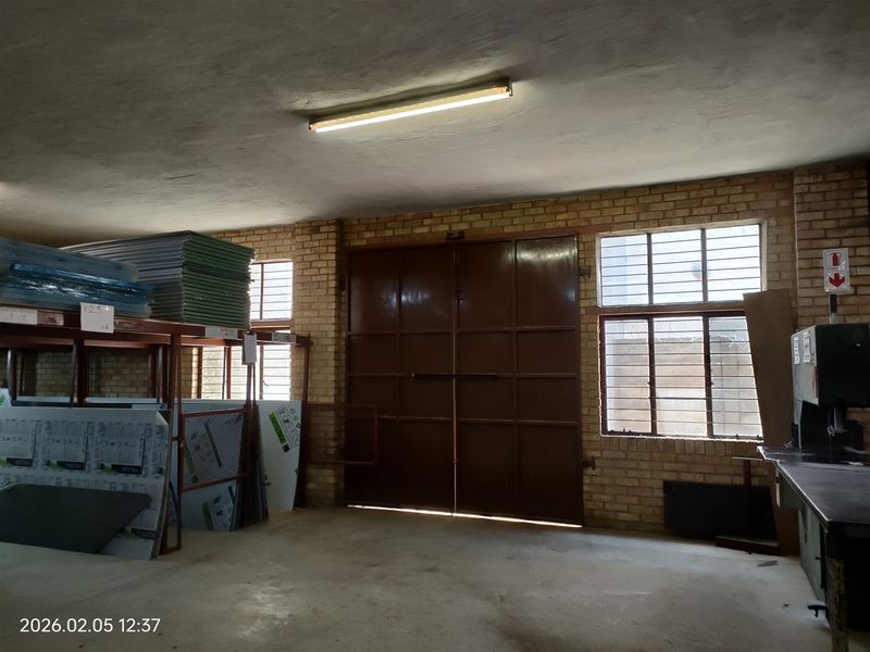Industrial utility space featuring exposed brick walls, concrete flooring, large double access doors, and secure window bars, suitable for storage or workshop use.