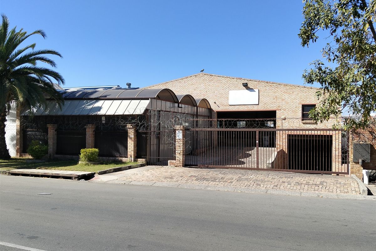 Front exterior view of an industrial property featuring a brick facade, secure sliding gate, razor wire fencing, and a curved polycarbonate awning over the entrance.