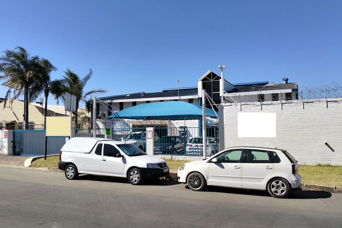 Front exterior view of the industrial property featuring a secure gated entrance, brick boundary wall with razor wire, and a shaded parking area.