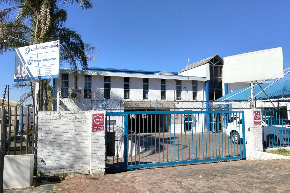 Front exterior view of an industrial property featuring a secure gated entrance, paved driveway, and prominent business signage.