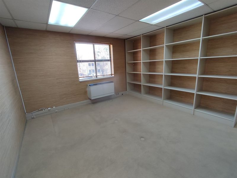 A vacant office space featuring built-in shelving, wood-look wall paneling, carpeted flooring, and a functional HVAC unit under the window.