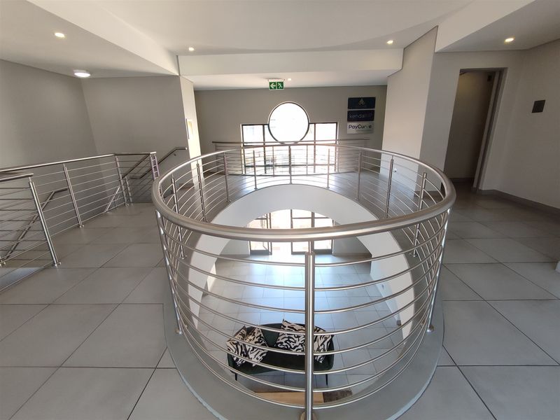 A modern office building hallway featuring a curved mezzanine landing with stainless steel balustrades, tiled flooring, and recessed lighting.