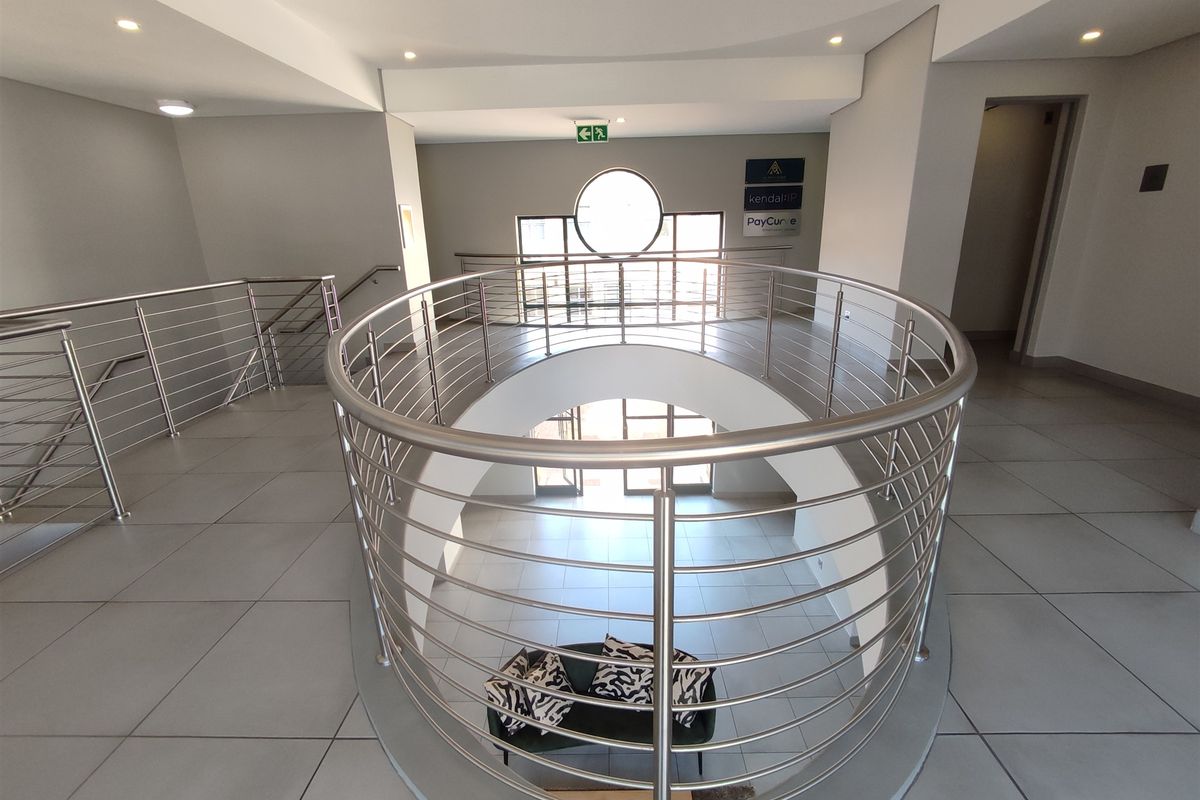 A modern office building hallway featuring a curved mezzanine landing with stainless steel balustrades, tiled flooring, and recessed lighting.