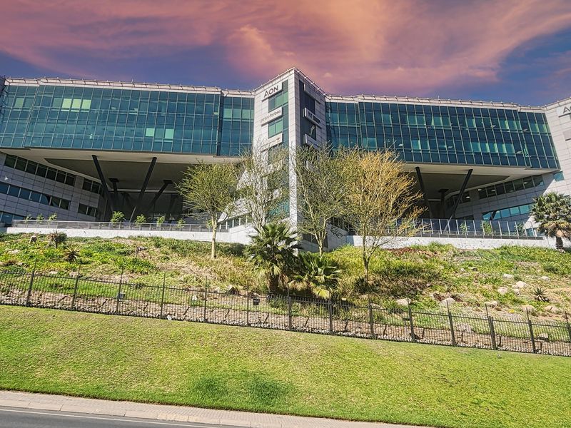 Modern multi-story office building featuring a glass curtain wall facade, prominent steel structural supports, and a well-maintained landscaped embankment with perimeter fencing.