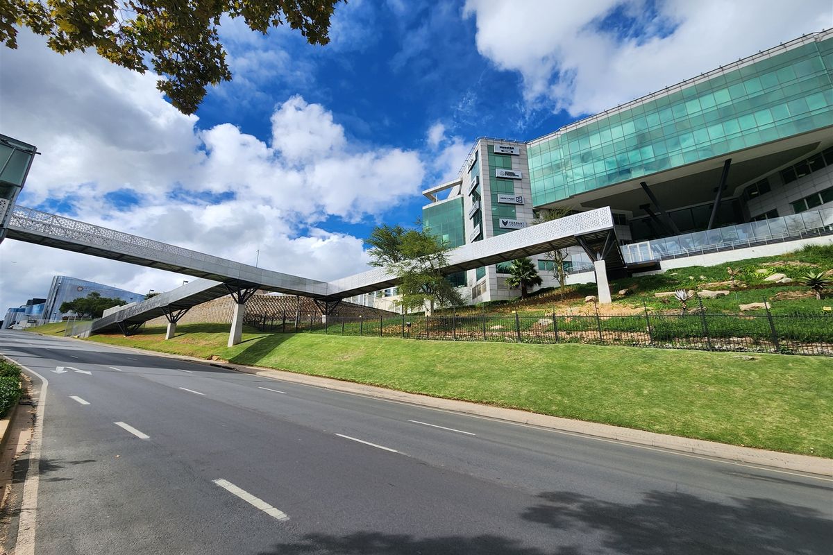 Modern commercial office building featuring a glass facade and an elevated pedestrian skybridge, situated above a landscaped roadside embankment.