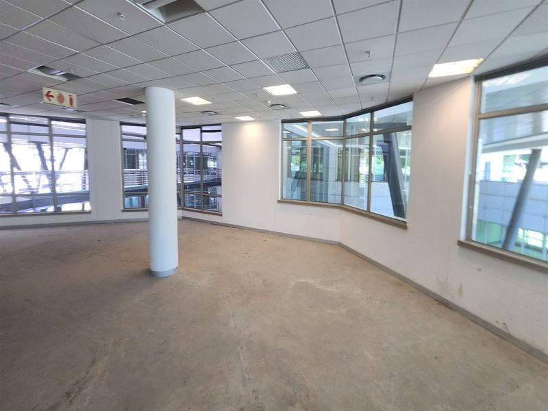 An empty, open-plan office space featuring large windows, a suspended ceiling with recessed lighting, and a central structural column.