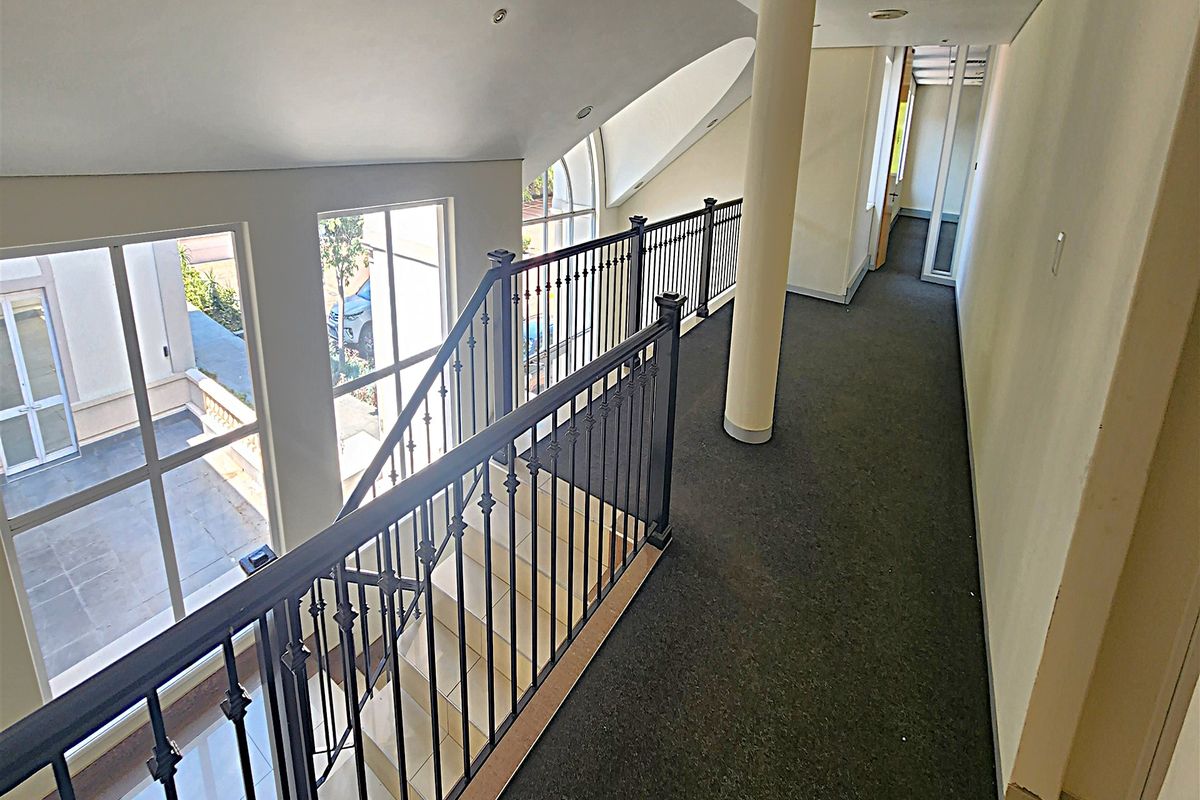 A spacious office hallway featuring a wrought iron balustrade overlooking the staircase, large windows for natural light, and a central structural column.