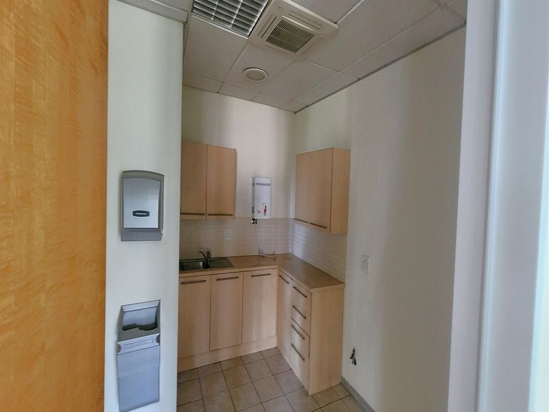 A compact office kitchenette featuring light wood cabinetry, a stainless steel sink, tiled backsplash, and integrated ceiling air conditioning.
