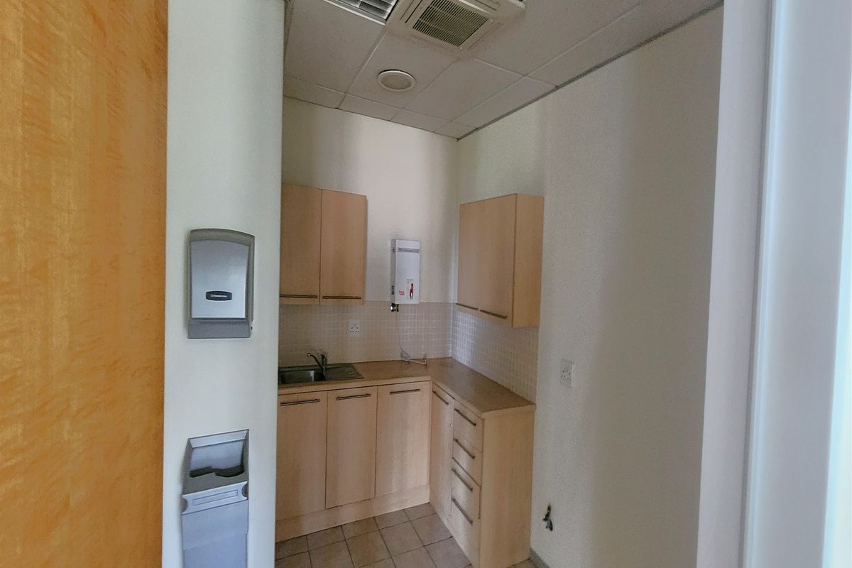 A compact office kitchenette featuring light wood cabinetry, a stainless steel sink, tiled backsplash, and integrated ceiling air conditioning.