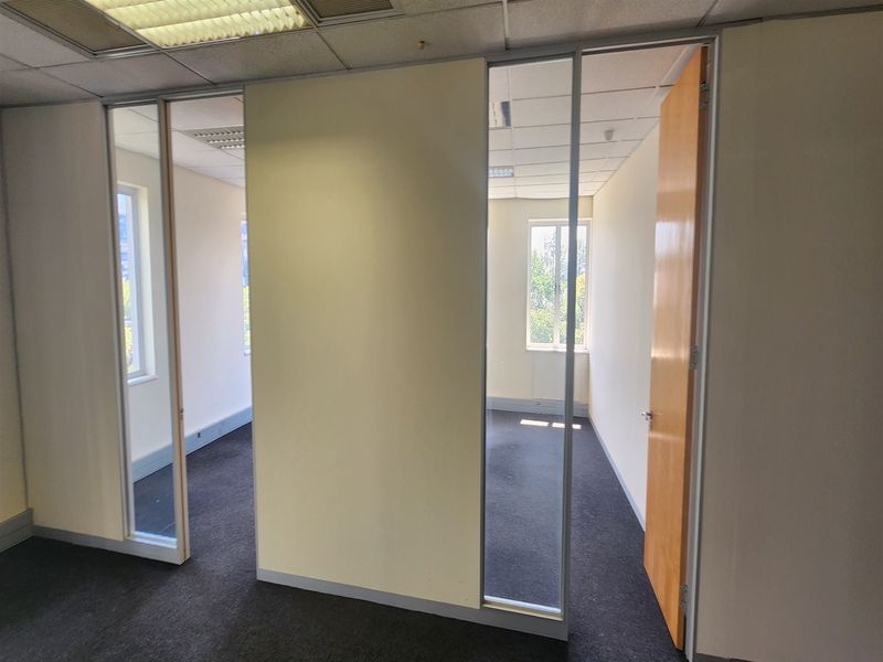 An empty office space featuring partition walls with glass panel doors, suspended ceilings with recessed lighting, and dark carpet flooring.