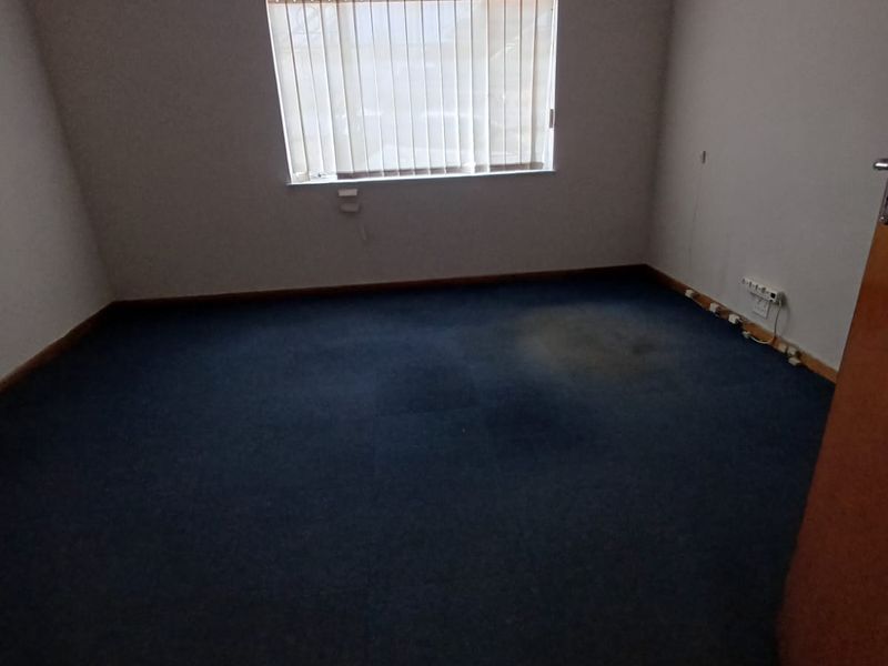 An empty office space featuring blue carpet flooring, a wall-mounted air conditioning unit, and vertical window blinds.