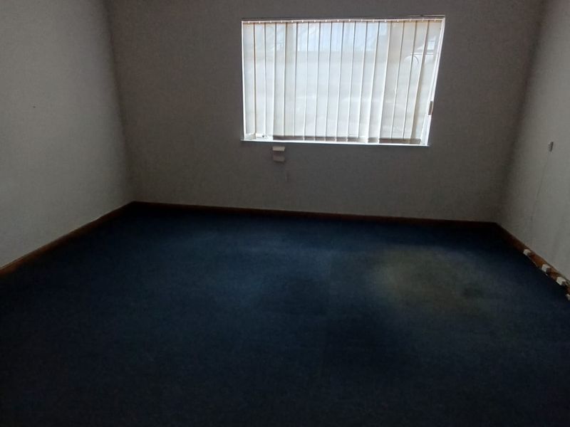 An empty office space featuring dark carpet flooring, a wall-mounted air conditioning unit, and vertical window blinds.