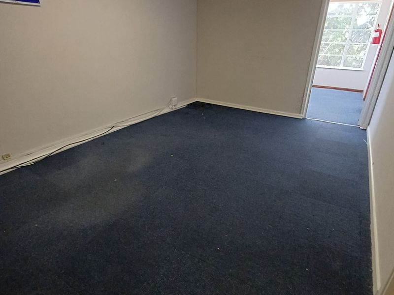 An empty office space featuring blue carpet flooring, a wall-mounted air conditioning unit, and overhead fluorescent lighting, with a doorway leading to an adjacent room.