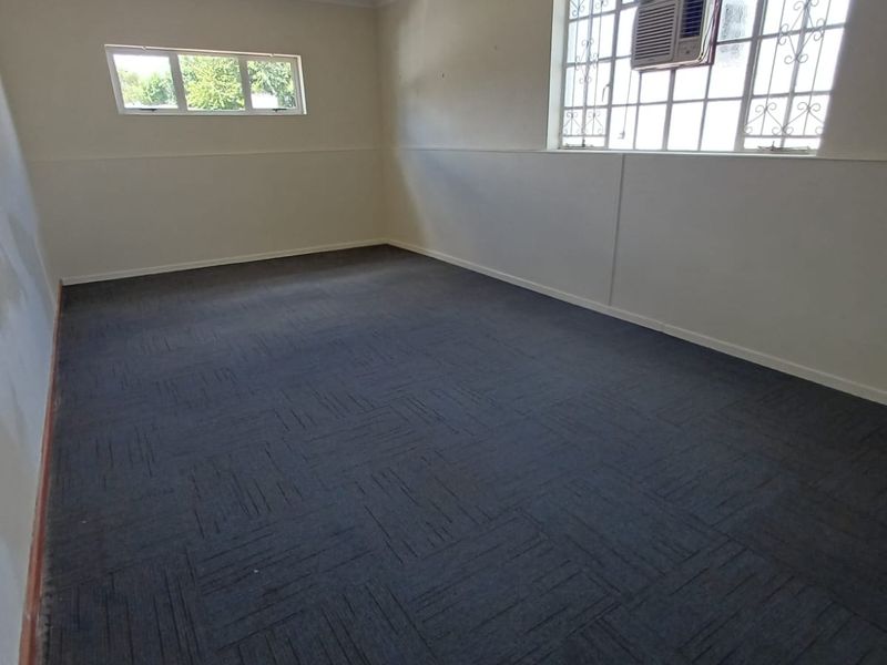 An empty office space featuring carpet tiles, window security bars, and a wall-mounted air conditioning unit, suitable for a professional workspace.