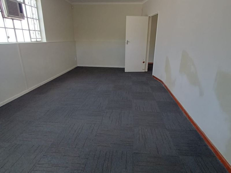An empty office space featuring carpet tiles, a window-mounted air conditioner, and fluorescent lighting, requiring minor cosmetic repairs to walls and doors.