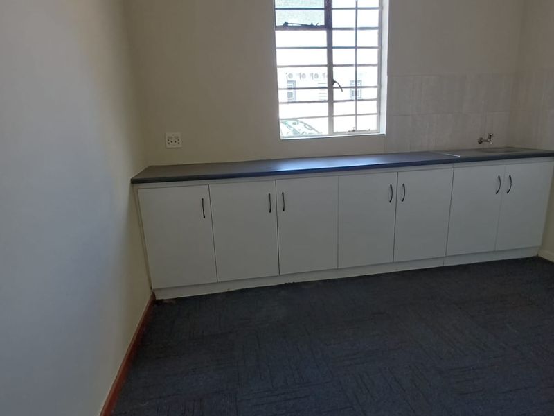 A clean, empty kitchenette area featuring white built-in cabinetry, a dark laminate countertop, a sink, and a window with security bars.