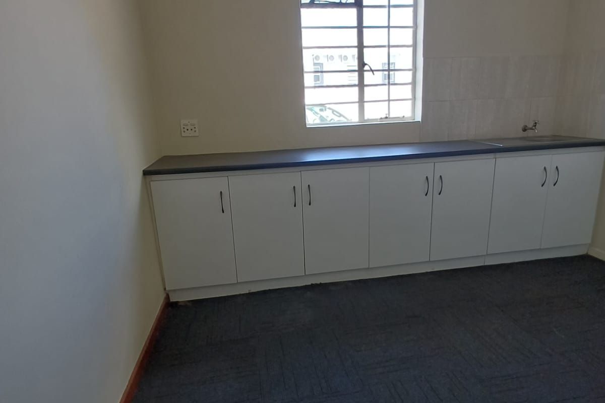 A clean, empty kitchenette area featuring white built-in cabinetry, a dark laminate countertop, a sink, and a window with security bars.