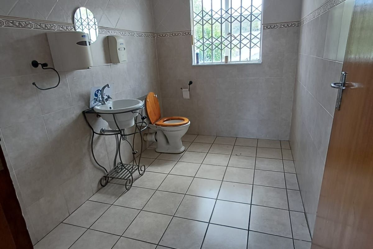 A functional bathroom featuring tiled walls and flooring, a pedestal washbasin, and a toilet, with a security gate installed on the window.