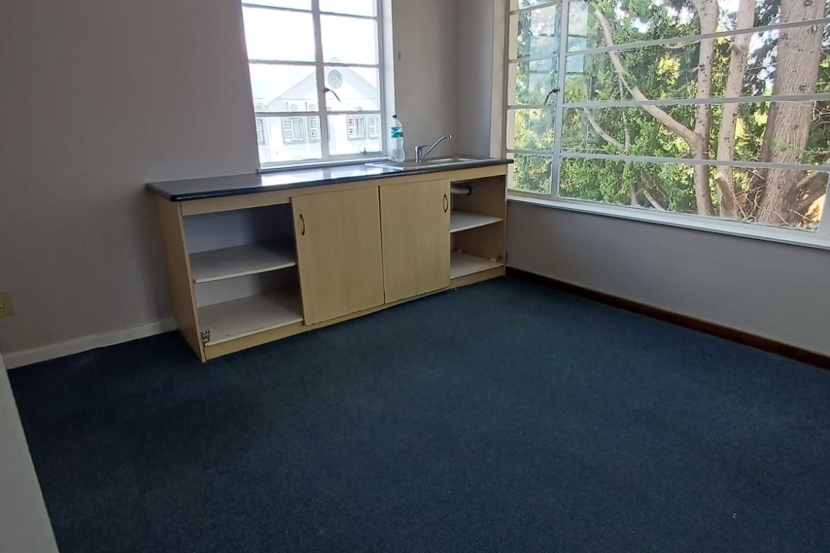 A small, empty kitchen area featuring built-in cabinetry with a sink, large windows providing natural light, and dark carpet flooring.