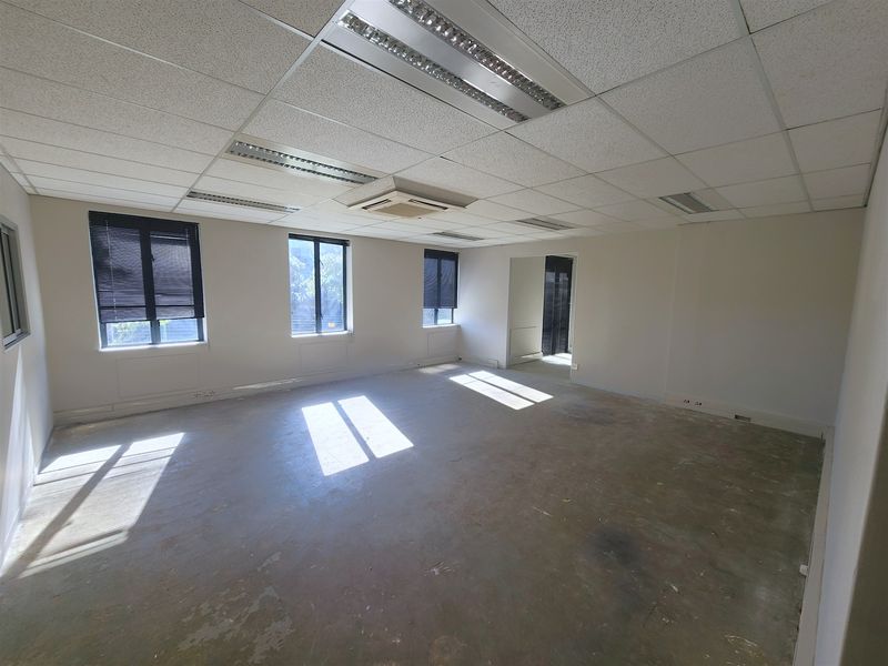An empty, open-plan office space featuring a suspended ceiling, recessed lighting, integrated air conditioning, and large windows with venetian blinds.